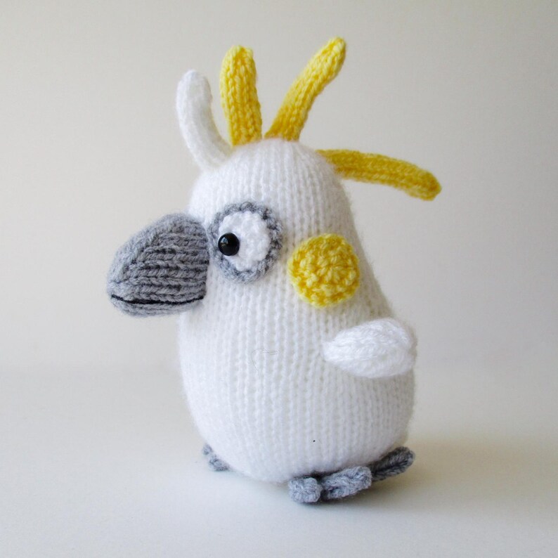 May include: A knitted cockatoo toy featuring a white body, a grey beak, and yellow crest feathers. The bird has a round yellow patch on its cheek, black bead eyes, and small grey feet. The texture is consistent with yarn knitting throughout.