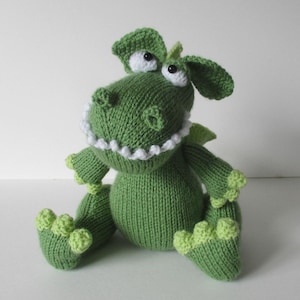 May include: A green knitted dragon plush toy featuring a round body, large white eyes with black pupils, and a wide mouth with small white teeth. The creature has light green claws on its hands and feet, and small wings on its back.