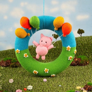 Puede incluir: Una corona de punto azul y verde, adornada con flores blancas. Un cerdito rosa sostiene globos de colores. La corona está suspendida sobre un fondo de hierba y cielo azul.