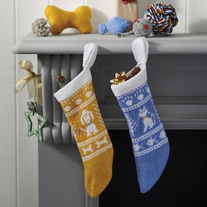 Puede incluir: Dos calcetines de Navidad de punto, uno amarillo mostaza y otro azul, cada uno con un diseño de perro blanco. Los calcetines cuelgan de una repisa de chimenea, con juguetes y adornos para perros encima. El calcetín amarillo tiene un patrón de perro y hueso.