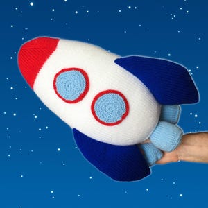May include: A knitted rocket ship toy featuring a white body, a red nose cone, and dark blue fins. The rocket has two circular light blue windows outlined in red, and light blue thrusters at the base, set against a dark blue starry background.