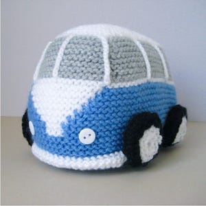 May include: A hand-knitted toy van featuring a blue and white body with a grey roof section. The vehicle has white yarn detailing for windows, two white circular buttons for headlights, and four black and white wheels attached to the sides.