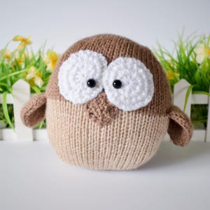 May include: A round, knitted owl toy featuring a two-tone brown body with a darker brown upper section and a lighter beige lower section. It has two large, circular white crocheted eyes with black plastic pupils and a small, triangular brown beak.