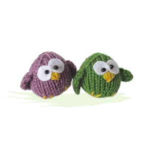May include: Two knitted bird figurines. One is purple, the other is green. Both have large white eyes with black pupils, yellow beaks, and yellow feet. The birds are small and round, with small wings.