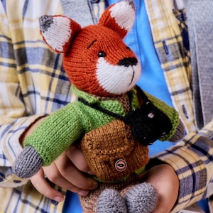 May include: A knitted fox plush toy with orange and white fur, wearing a green sweater, a brown satchel with a single button, and a black camera strap. The fox has grey paws and pointed ears with white tips, crafted in a soft yarn texture.