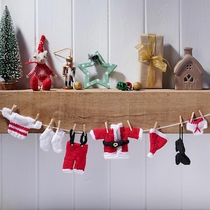 May include: A Christmas-themed scene with miniature knitted garments strung up with pegs. The items include a sweater, socks, dungarees, a Santa suit, a hat, and underwear. A small Christmas tree, a mouse figure, and other festive decorations are on a wooden shelf.