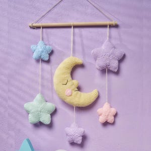 May include: A handcrafted nursery mobile with a yellow crescent moon and five knitted stars in pastel shades of blue, green, lilac, and pink. The mobile is suspended from a wooden dowel using string.
