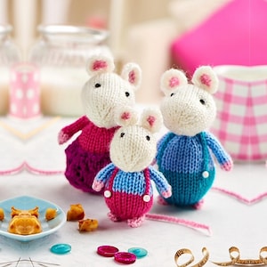 May include: Three handmade knitted mice toys. The mice have white bodies, pink ears and noses, and are wearing blue, pink, and purple outfits. The toys are on a white surface with small decorative items.