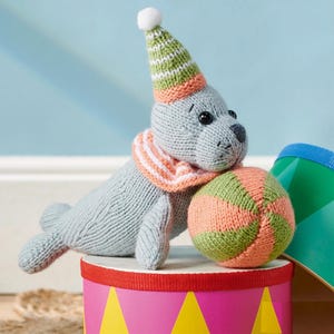 May include: A gray knitted seal toy wearing a striped green and orange hat and scarf, with a matching ball. The seal is resting on a pink and yellow drum.