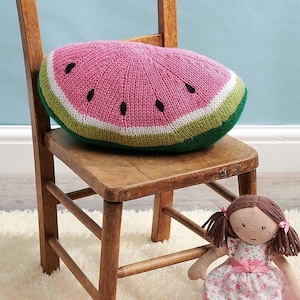 May include: A pink and green knitted watermelon cushion on a wooden chair. The cushion is shaped like a slice of watermelon and has black seeds.