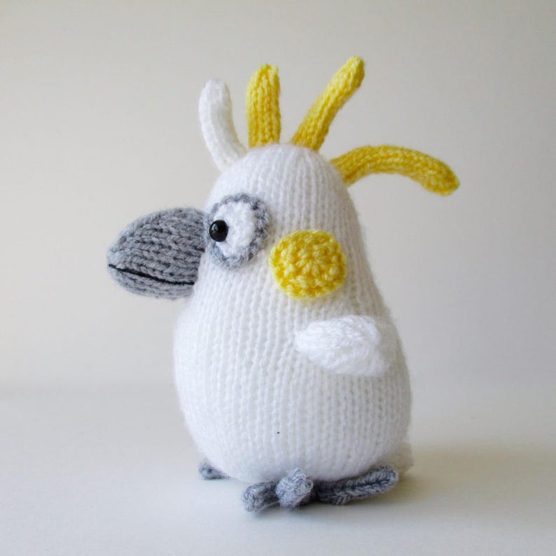 May include: A hand-knitted cockatoo plush toy featuring a white body, a grey beak, and grey feet. The bird has a yellow crest on its head, a yellow circular patch on its cheek, and a black eye surrounded by a grey ring, all crafted from yarn.
