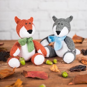 May include: Two knitted animal dolls: a fox and a wolf. The fox is orange, white, and black, wearing a green scarf. The wolf is gray, white, and black, with a blue scarf. Both dolls have acorn accessories and are on a wooden surface.