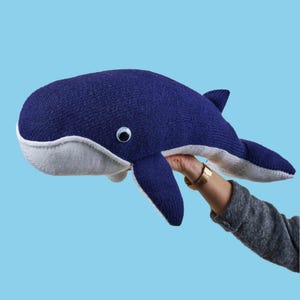 May include: A dark blue knitted whale plush toy with a white underbelly and a single googly eye. The whale features a rounded head, two side fins, and a tail fin, constructed from a textured knit fabric with visible stitching along the edges.