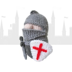 Puede incluir: Muñeco de caballero tejido, con casco gris, cubrecara y cuerpo. El muñeco sostiene una espada gris y un escudo blanco con una cruz roja. El fondo presenta una silueta de castillo.