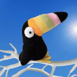 May include: A knitted toucan bird with a black body, a colorful striped beak, and a white eye with a black center. The toucan is perched on a white branch against a bright blue sky.