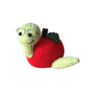 May include: A hand-knitted soft toy shaped like a snail with a light green head and body, wearing small silver-toned wire-rimmed glasses. The shell is a bright red apple with a dark brown stem and a small dark green leaf attached to the top.