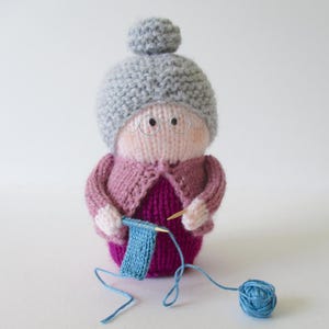 May include: A knitted doll featuring a grey bun hairstyle, wire-rimmed glasses, a pink cardigan, and a dark magenta dress. The doll holds a small blue knitted square on wooden needles, with a matching blue ball of yarn connected by a thread.