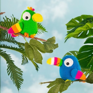 Peut inclure: Deux ornements d'oiseaux tricotés perchés sur des branches. L'un est vert avec un bec jaune et des accents d'ailes arc-en-ciel. L'autre est bleu avec un bec blanc, rose et jaune et des accents d'ailes arc-en-ciel. Le fond est un ciel bleu clair avec des feuilles vertes.