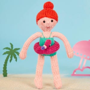 Peut inclure: Une poupée en tricot portant un maillot de bain vert et un flotteur rose en forme de flamant rose. La poupée a les cheveux roux et le visage rose. La poupée est debout sur une surface sablonneuse.