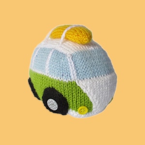 Könnte beinhalten: Ein handgestrickter Spielzeugbus in Weiß, Hellblau und Grün, mit schwarzen Rädern und einem gelben Akzent oben. Der Bus hat Knöpfe und steht vor einem gelben Hintergrund.
