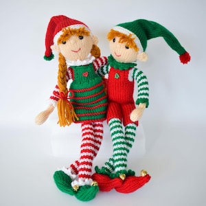 May include: Two handmade knitted dolls featuring elf-like designs. One doll wears a red hat and green striped dress with a red heart button. The other wears a green hat and red overalls with a green tree button. Both have striped legs and gold bells.