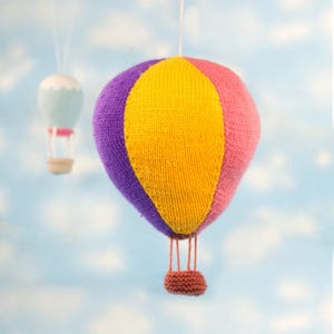 May include: A knitted hot air balloon toy with a purple, yellow and pink colour scheme. The balloon is hanging from a string and has a small basket.
