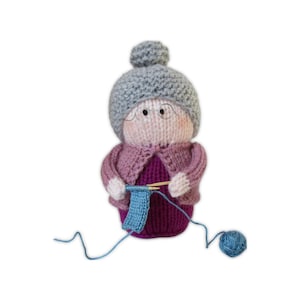 Puede incluir: Muñeca tejida a mano de una abuela con un gorro gris con pompón, gafas y un cárdigan rosa. La muñeca está tejiendo con hilo azul y tiene una madeja de hilo azul cerca. La muñeca lleva un vestido morado.