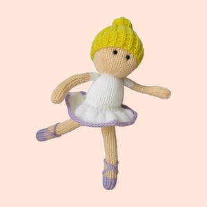 May include: A knitted ballerina doll with a yellow cap, tan arms and legs, and a white dress with a lavender trim. The doll is wearing lavender ballet slippers and is posed mid-leap.