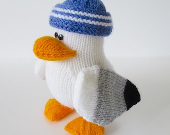 May include: A knitted seagull toy featuring a white body, orange beak and feet, and a blue knit cap with white stripes. The wings are grey with black tips, and the eyes are small black beads. The toy is crafted with a visible knit texture.