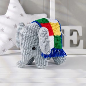 May include: A gray knitted elephant toy with large ears and a small black button eye. The elephant is wearing a colorful patchwork blanket with blue fringe. A white star-shaped pillow is in the background.