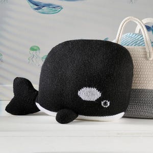 Puede incluir: Un peluche de orca tejido en blanco y negro con vientre y marcas oculares blancas. La ballena tiene una pequeña aleta y cola. Una cesta decorativa con asa de cuerda está en el fondo.
