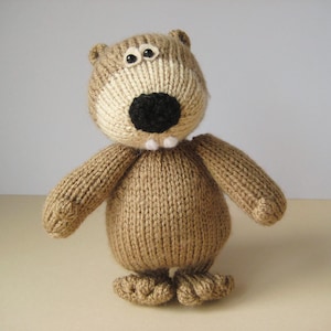 May include: A hand-knitted stuffed animal featuring a light brown textured body, a round head with two small white eyes with black pupils, a large black nose, and two small white teeth protruding from the bottom of the muzzle area.
