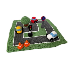 May include: A knitted play mat with a road design, featuring toy cars, traffic cones, a tree, and a traffic light. The mat is green with gray roads and white borders. The toy cars are red, white, and yellow.