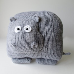 May include: A gray knitted stuffed animal shaped like a hippopotamus. The toy features a rounded body, four short legs, and a small tail. It has two white circular eye patches with black beads for pupils, small ears, and a snout with nostrils.