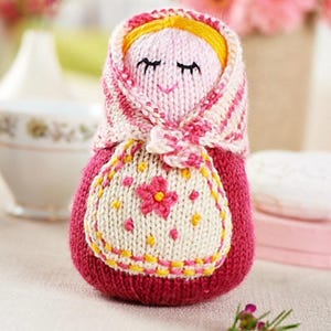May include: A handmade knitted doll with a red body, a white apron with a pink flower, and a pink and white striped head covering. The doll has yellow hair and closed eyes, and is a charming, handcrafted item.