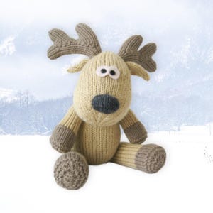 May include: A knitted reindeer plush toy with a light tan body, brown antlers, and gray accents on the nose and feet. The reindeer has large, round eyes and is sitting down. The background is a winter scene.