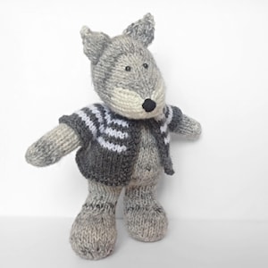 May include: A hand-knitted stuffed animal shaped like a wolf with grey and cream yarn. The wolf features a black nose, dark eyes, and pointed ears. It is wearing a dark grey and white striped knitted cardigan that covers its torso and arms.