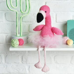 May include: A pink knitted flamingo doll with a black beak, white eye accents, and a fluffy pink tutu. The flamingo has long, thin legs and is sitting on a white shelf. The background includes a green cactus-shaped light and yarn balls.