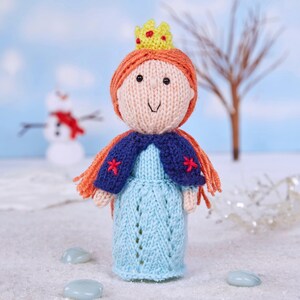 Peut inclure: Poupée princesse tricotée à la main, avec des cheveux en fil orange, une couronne jaune et une robe bleue. La poupée porte une cape bleue avec des étoiles rouges. Un bonhomme de neige est en arrière-plan.