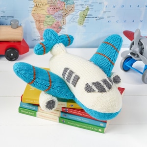 May include: A white knitted airplane toy with blue wings and orange stripes. The toy is sitting on a stack of books.