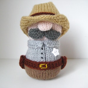 May include: A hand-knitted doll shaped like a sheriff, featuring a tan hat, a pink nose, a dark gray mustache, a light gray shirt with black buttons, a white star-shaped button badge, and a brown belt with a yellow embroidered buckle.