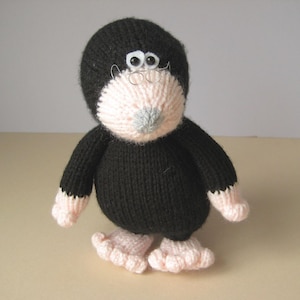 May include: A knitted plush toy shaped like a mole with a black body, head, and arms. It features a pale pink face, hands, and feet, with a small grey nose. The toy has two white plastic eyes and a pair of thin, circular wire-rimmed glasses.