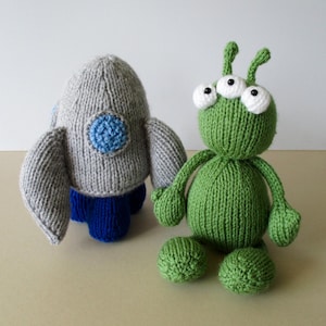 May include: A hand-knitted gray rocket ship toy with a blue circular window and blue base, positioned next to a green knitted alien toy featuring three white eyes with black pupils and two antennae on top of its round head.