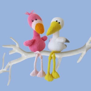 Puede incluir: Dos muñecos de aves tejidos sentados en una rama blanca. El ave de la izquierda es rosa con pico naranja y patas rosa claro. El ave de la derecha es blanca con pico amarillo, puntas de alas grises y patas amarillas. Ojos de cuentas negras.
