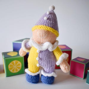 May include: A hand-knitted doll featuring a pointed purple hat with white pom-poms, a peach-colored face, and a two-toned yellow and purple body. The doll wears a white ruffled collar and cuffs, with blue knitted shoes, standing among colorful blocks.