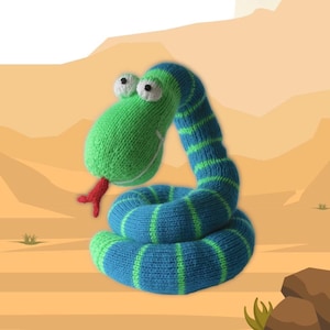 Op de afbeelding: Een gebreid slangenknuffel in groen en blauw, met grote witte ogen en een rode tong. De slang is opgerold, met groene en blauwe strepen. De achtergrond is een woestijnlandschap met zand en enkele planten.