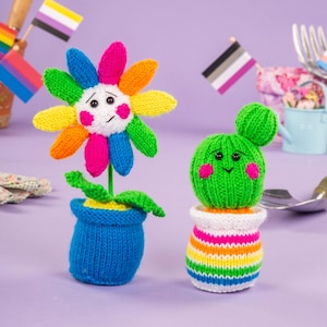 Könnte beinhalten: Zwei gestrickte Pflanzenfiguren: eine bunte Blume und ein grüner Kaktus. Die Blume hat Regenbogenblütenblätter und einen blauen Topf. Der Kaktus hat einen grünen Körper, rosa Wangen und einen gestreiften Topf. Flaggen mit LGBTQ+-Symbolen sind im Hintergrund.