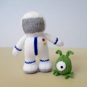 May include: A hand-knitted white astronaut doll with a grey visor, blue trim, and two small red and yellow buttons on the chest, accompanied by a small green knitted alien creature with one large white eye and two antennae on its head.