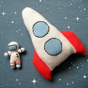 May include: A white knitted rocket ship plush toy with two light blue circular windows and bright red fins, accompanied by a small white knitted astronaut doll featuring a light blue visor and black and red accent details on the suit.