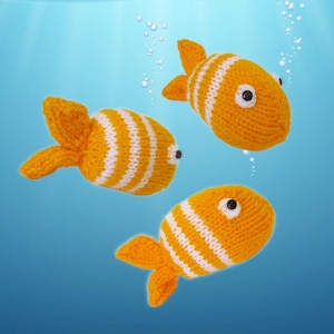 May include: Three knitted orange fish with white stripes and black eyes float against a blue background. The fish have orange fins and tails, and appear to be handmade. Bubbles are scattered around the fish.